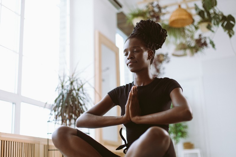 Femme noire yeux clos, assise en tailleur et mains jointes, en profonde méditation pour explorer les différents niveaux de conscience de son éveil spirituel.